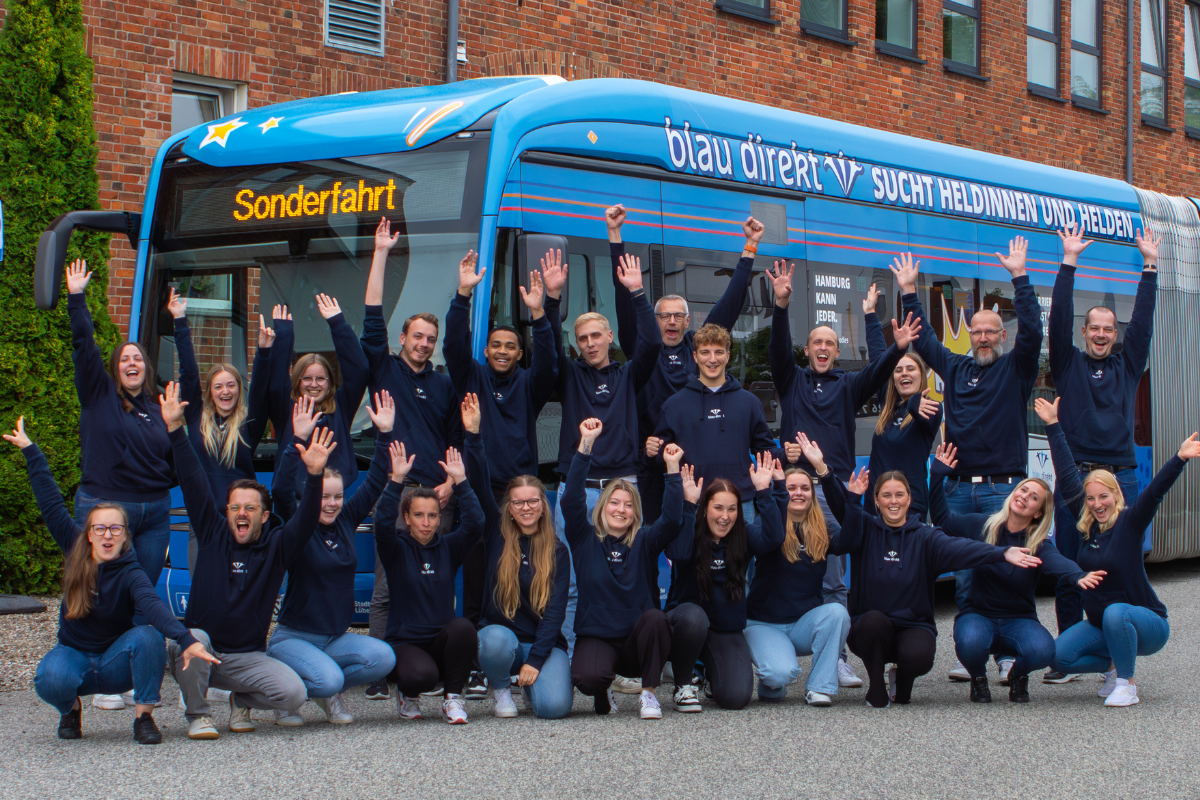 Eine Gruppe von Frauen und Männer trägen blau direkt Hoodies und lächeln mit den Ärmeln hoch vor einem Bus mit einer blau direkt Werbung. Die Werbung zeigt ,dass blau direkt Heldinnen und Helden sucht.
