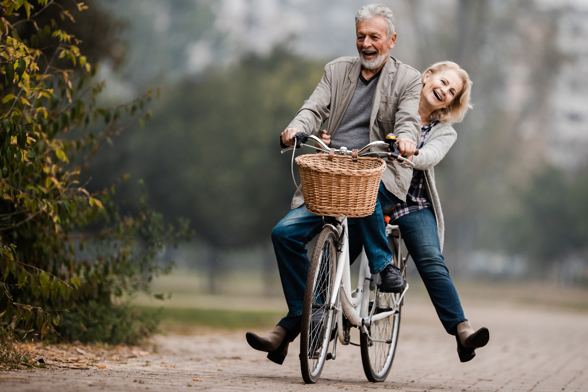Man sieht ein lachendes Rentner Paar auf dem Fahrrad. Ganz links sind Bäume zu sehen.