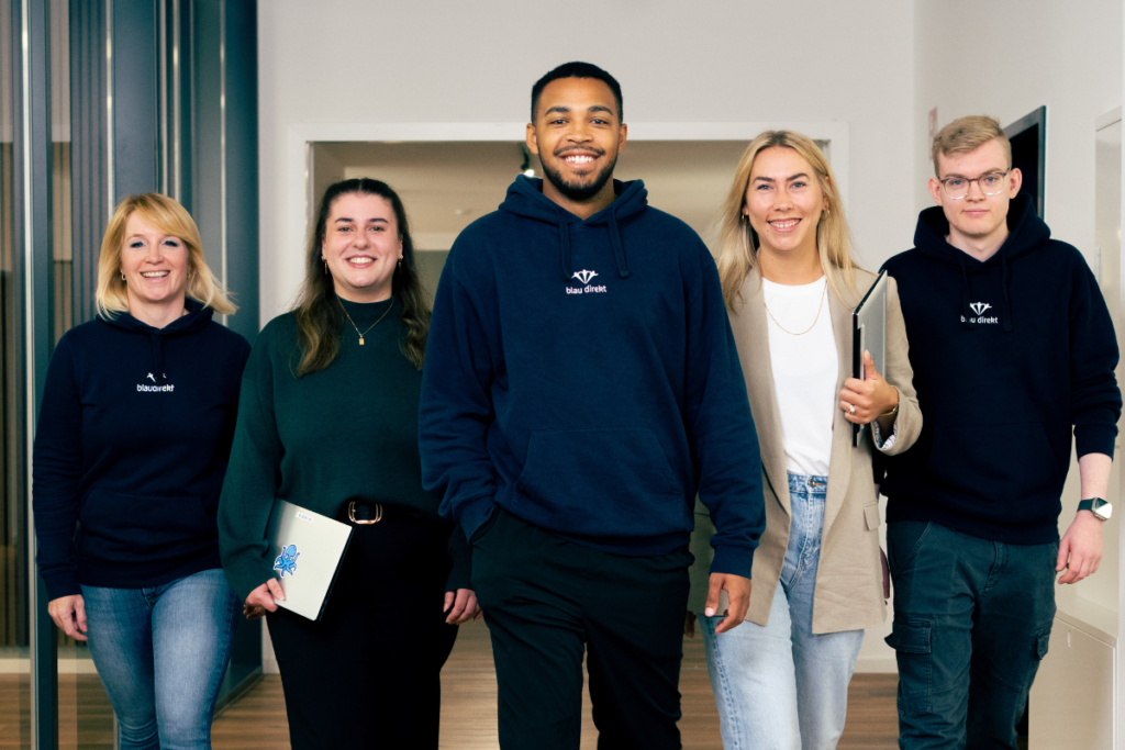 Es sind drei Frauen und zwei Männer zu sehen. Alle lächeln laufen in Richtung Kamera. Drei von ihnen tragen blau direkt Hoodies.