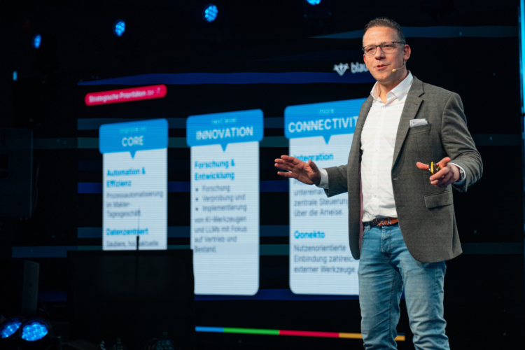 Ein Mann hält eine Rede auf der Bühne mit einer offenen Haltung. Hinter ihm ist eine Präsentation mit den Kategorien Core, Innovation, Connectivity zu sehen,