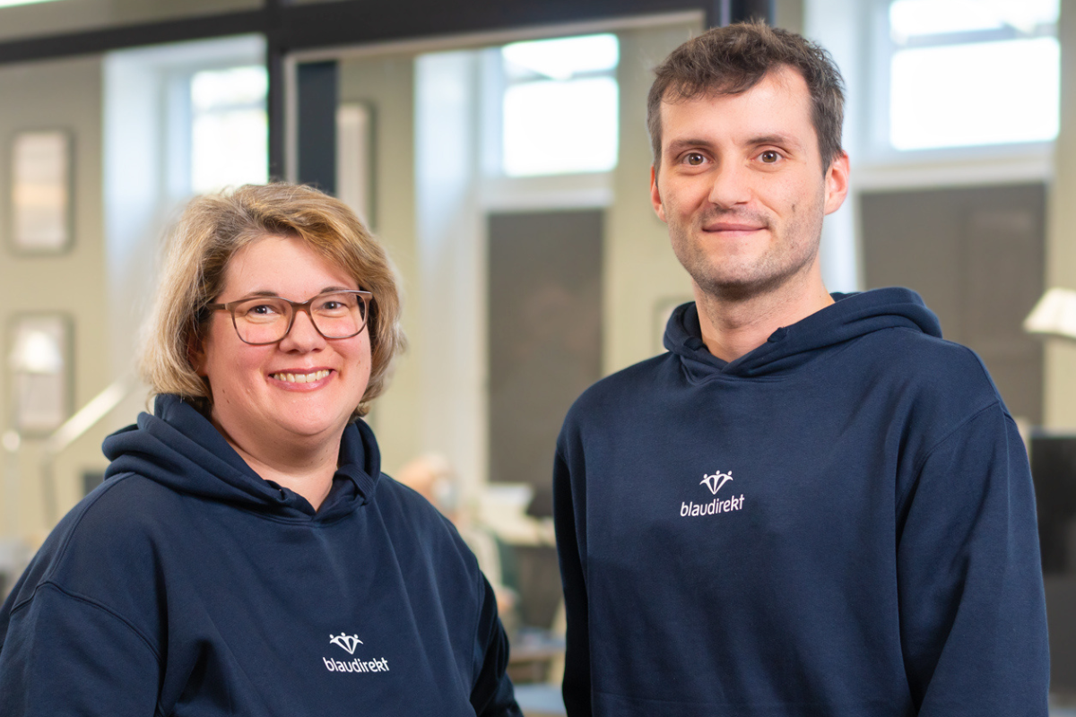 Eine Frau mit Brille und ein junger Mann jeweils im blau direkt Hoodie lächeln in die Kamera.