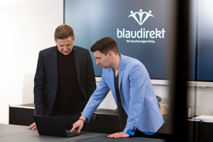 Zwei Männer stehen vor einem Screen mit dem blau direkt Logo. Die beiden Männer schauen gemeinsam auf einen Laptop.