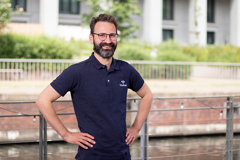 Im Vordergrund ist ein lächelnder Mann mit einer aufrichtigen Haltung zu sehen. Er trägt ein blau direkt Polo T-Shirt. Im Hintergrund sieht man ein Gebäude und Bäume.