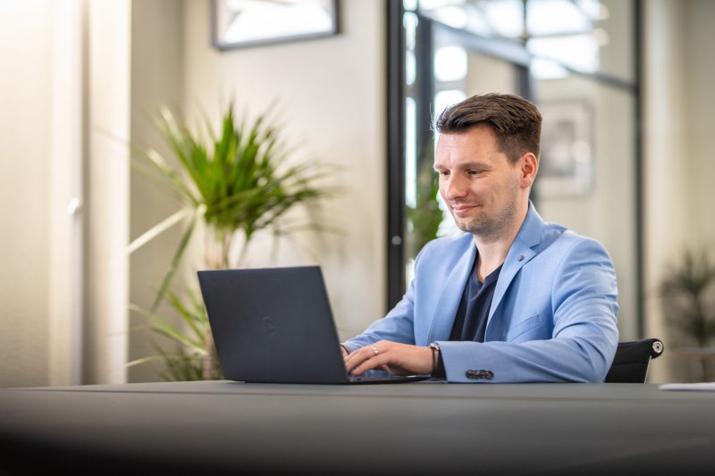 Auf den Bild ist ein Mann, der auf seinem Laptop am Schreibtisch arbeitet. Er befindet sich in einem Büro mit einer Pflanze im Hintergrund.